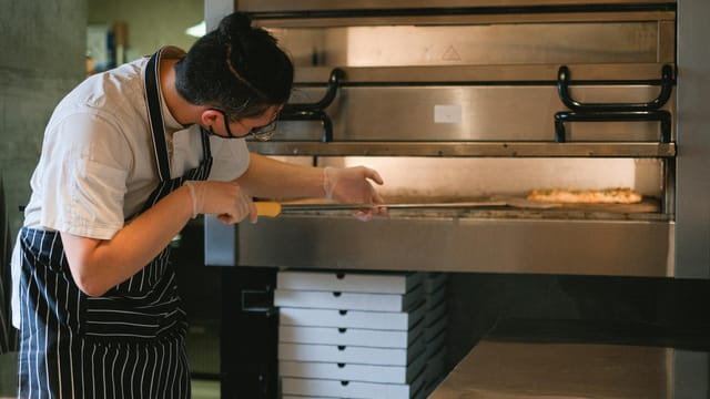 A chef in an apron prepares pizza in a commercial kitchen oven.