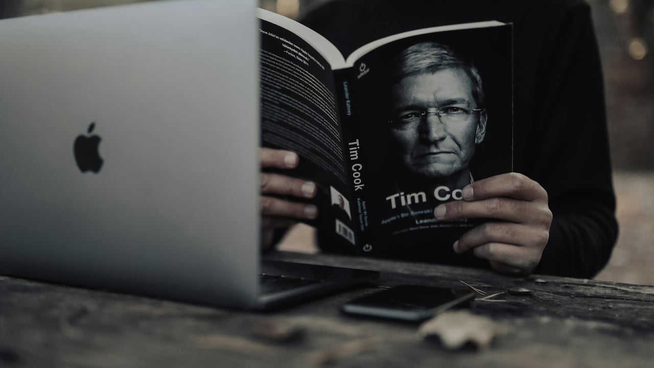 Unrecognizable ethnic male in warm clothes with opened laptop reading book and sitting outside at wooden table in autumn day