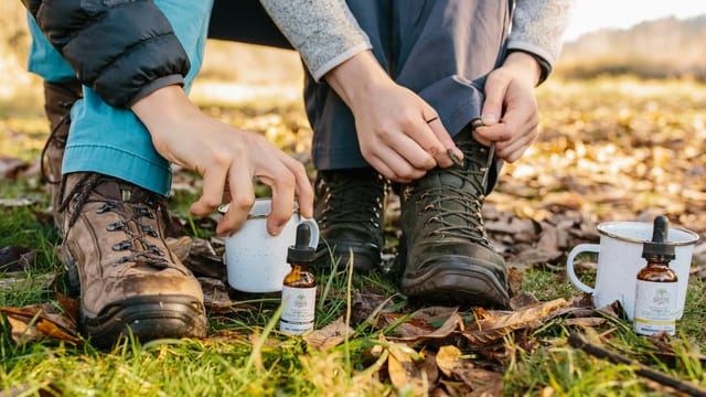 Close-up of hands adjusting hiking boots next to CBD oil and coffee cups outdoors.