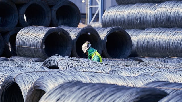 Construction worker in safety gear navigates large stacks of metal wire coils at an industrial site.