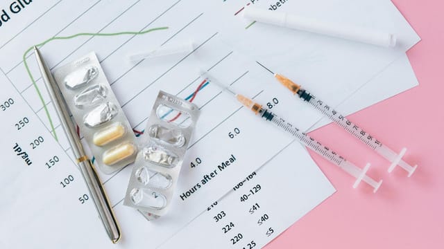 Flat lay of medicines, syringes, and a blood glucose chart on a pink surface, representing health management.