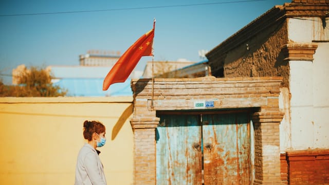 A woman in a face mask walks past a rustic building with a Chinese flag under a bright sky.