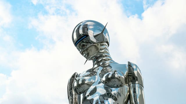 A sleek chrome robot sculpture stands against a bright blue sky background.