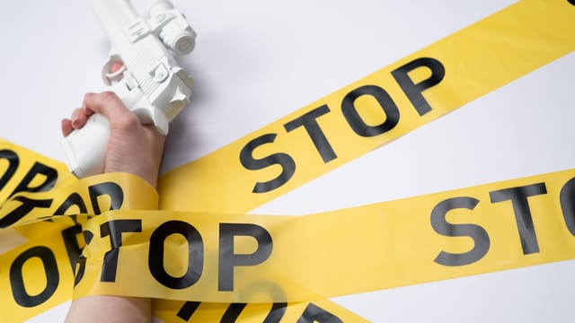 A white toy gun held by a hand wrapped in yellow stop tape on a white background, conveying a message of caution.