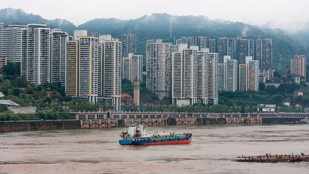 A cargo ship traverses a river with a backdrop of towering city buildings and misty mountains.