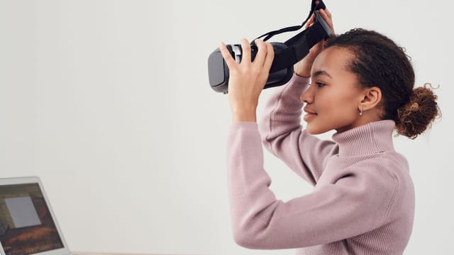 A woman puts on a VR headset in a modern room with a laptop nearby, exploring virtual environments.