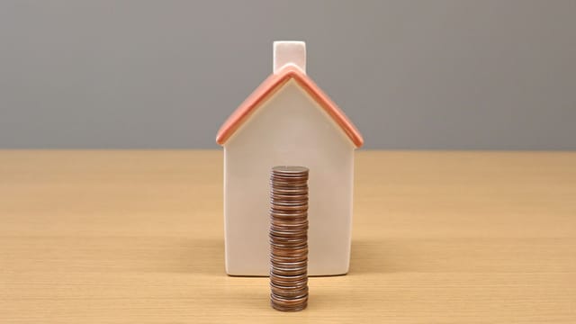 A conceptual still life image of stacked coins in front of a porcelain house, symbolizing savings and investment.