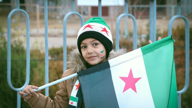 A young girl in Syria proudly holds the Syrian flag, symbolizing hope and resilience.