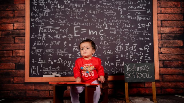 A baby in a classroom setting with a math-filled chalkboard background.