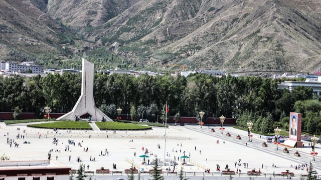 City square bustling with people, featuring a prominent monument and scenic mountain backdrop.