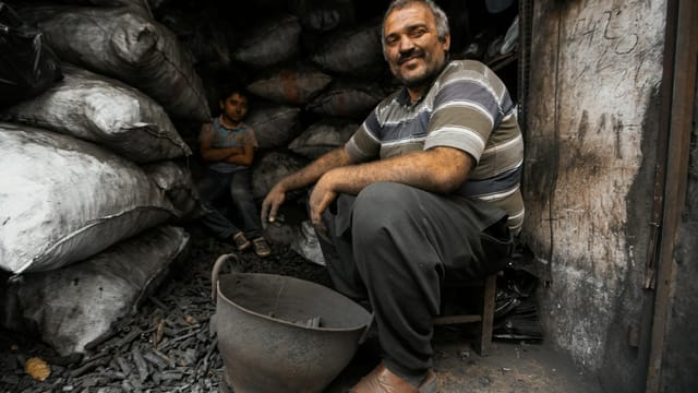 A charcoal worker smiles while sitting in a dimly lit workshop with bags of charcoal.