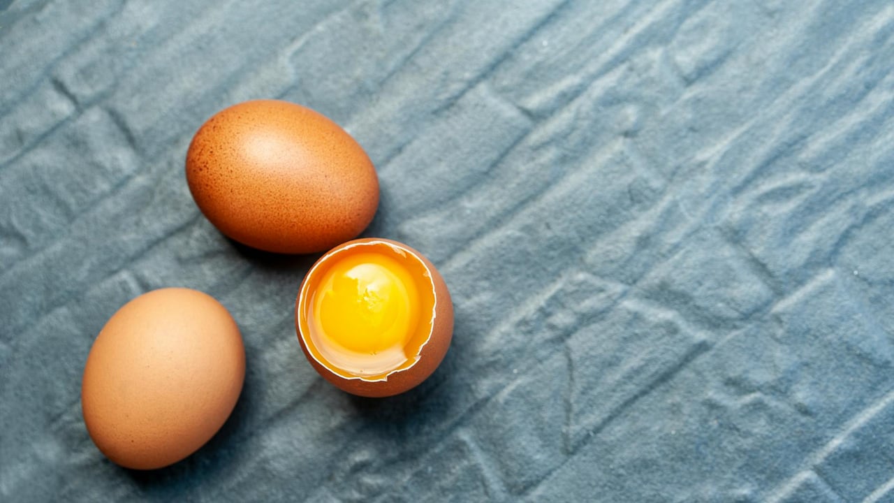 Close-up of three brown eggs on textured blue fabric; one egg is cracked open revealing the yolk.