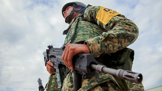 Close-up of a soldier in camouflage holding a rifle under a blue sky, representing military readiness in Mexico.