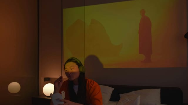 An Asian woman with headphones enjoys eating on bed watching a movie projection.