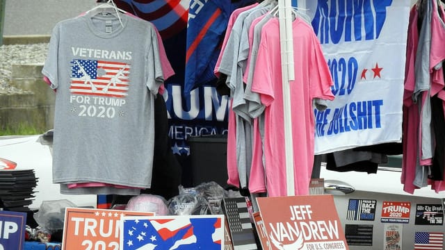 A stall displaying Trump 2020 merchandise including shirts and signs at an outdoor market.