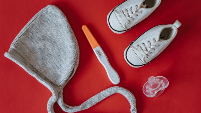 A flat lay image featuring baby shoes, a pregnancy test, and baby essentials on a red background.