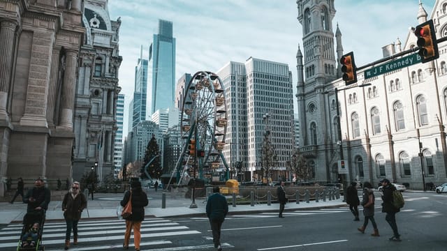 Urban street scene with people crossing, skyscrapers, Ferris wheel, and historical architecture under clear sky.