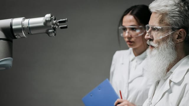 Two scientists wearing lab coats and goggles analyzing a robotic arm in a laboratory setting.