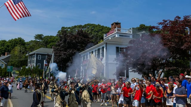 Crowded parade with reenactors in uniform firing rifles, surrounded by festive people waving flags on a sunny day.