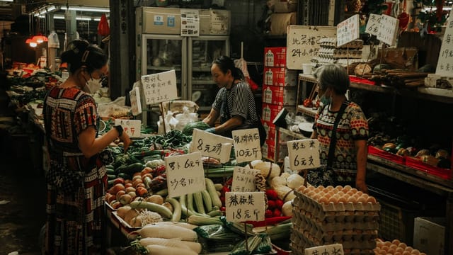 A lively market scene with women shopping for fresh veggies and eggs.