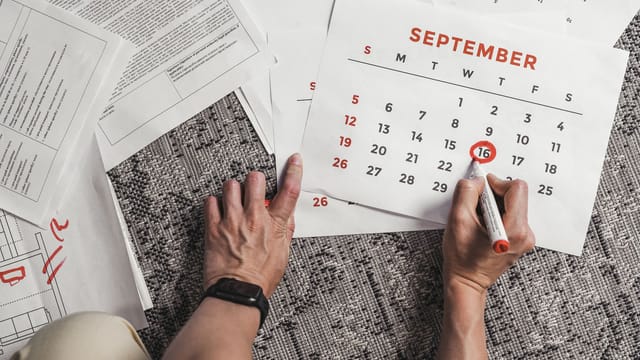 Hands holding a calendar with September dates encircled, surrounded by documents on a carpet.