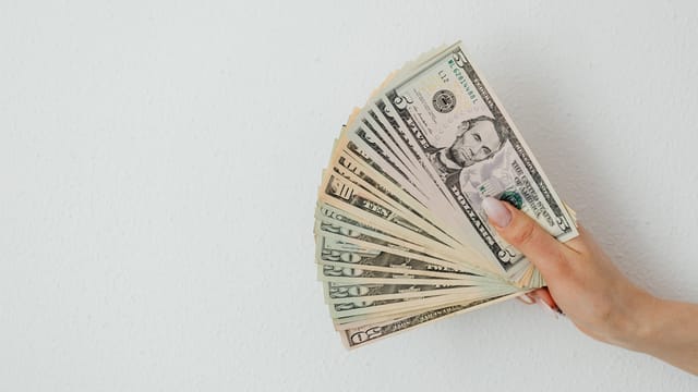 A hand holding an array of US dollar bills, fanned out against a plain white background.