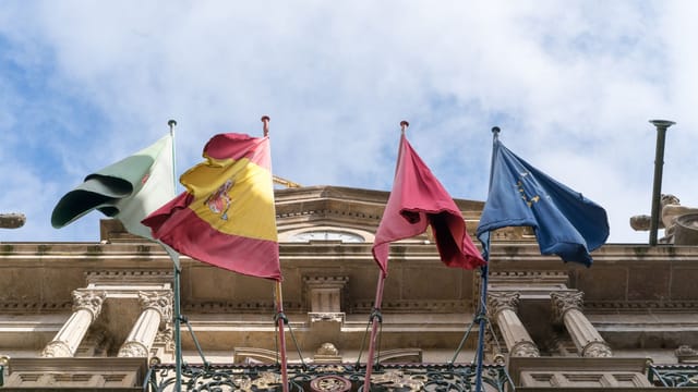 From below of Spanish flag and flags of Spanish provinces together with European Union flag waving on facade of magnificent stone building on sunny day