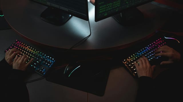 Two people typing on RGB keyboards with code on screens, indicating a cybersecurity environment.