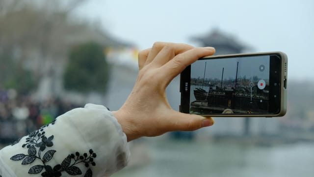 A hand holds a smartphone capturing a serene Chinese scene, blending technology and tradition.