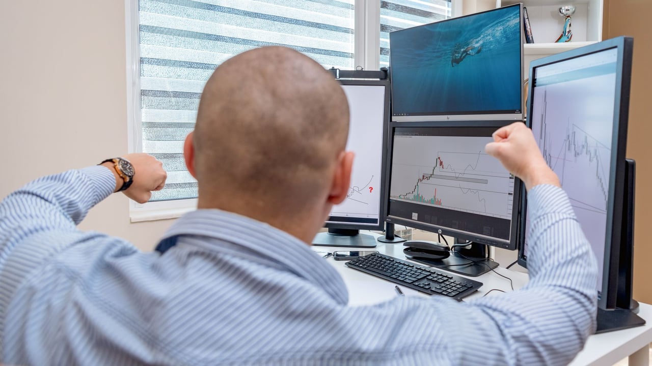 A man celebrates success at a multi-monitor workstation while analyzing stock charts.