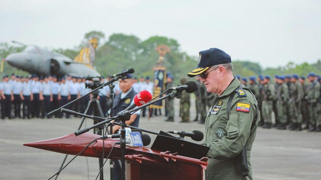 A military officer delivers a speech at an outdoor ceremony with an aircraft and soldiers in the background.