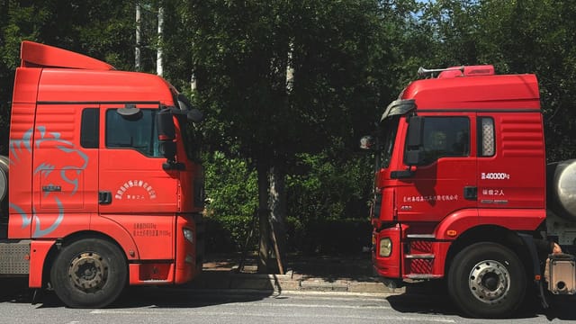 Two red trucks facing each other on a road surrounded by trees. Sunny day outdoors.
