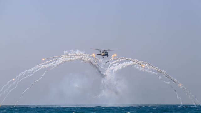 A helicopter executes a stunning aerial maneuver, releasing flares over the sea against a clear sky.