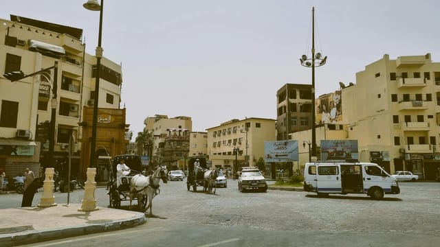 Busy intersection with horse-drawn carriages and vehicles in a Middle Eastern city.