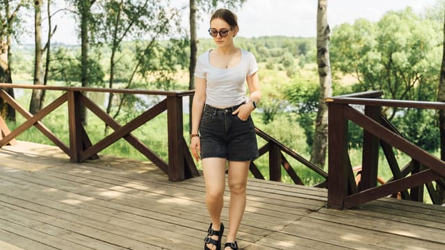 Woman in casual summer attire walks on a wooden deck amid lush green park.