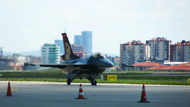 Fighter jet on airport runway with city skyline in the background, ready for takeoff.