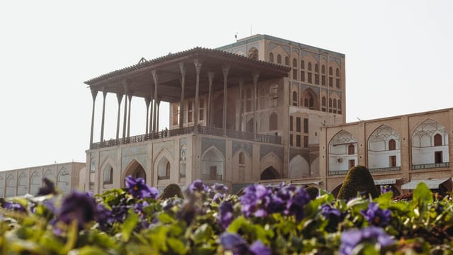 A beautiful view of Ali Qapu Palace in Esfahan, Iran with vibrant purple flowers in the foreground.