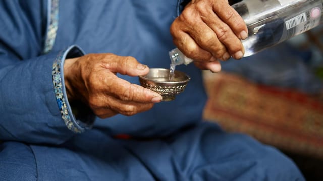 Crop anonymous senior male in national costume pouring vodka in Tibetan Singing Bowl sitting on carpet