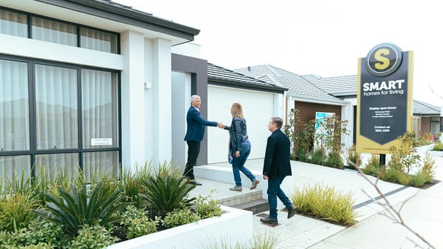 A realtor shakes hands with potential buyers outside a modern model home.