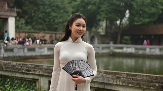 Beautiful young woman in traditional Vietnamese dress holding a fan by a historic pond.