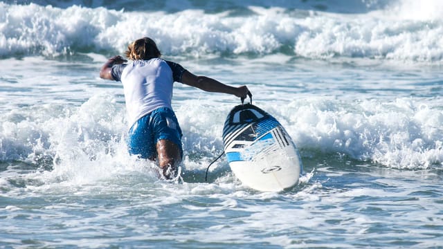 A surfer with a surfboard heads into the waves at Tel Aviv beach in bright daylight.