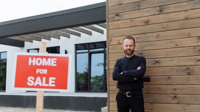 Real estate agent standing by a modern home with a 'Home for Sale' sign.