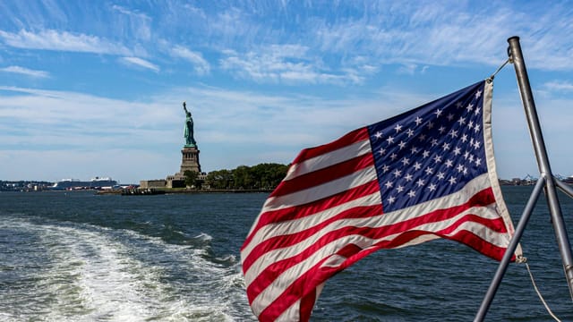 American flag waving with Statue of Liberty in the background, captured from a boat in New York Harbor.