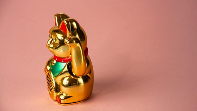 Gold Maneki Neko figurine, a Japanese lucky cat, displayed on a pastel pink background.