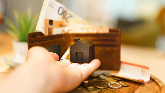 A hand holding a small house model with euro notes and coins nearby, illustrating real estate investment and finance.