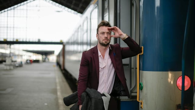 Thoughtful male passenger wearing formal outfit carrying warm coat with hand near head while standing near train with opened door on platform of railroad station during business trip