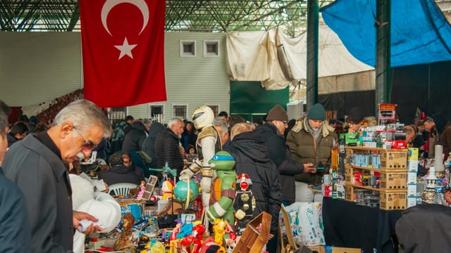 Busy flea market in Turkey with shoppers surrounded by antiques and toys under a Turkish flag.