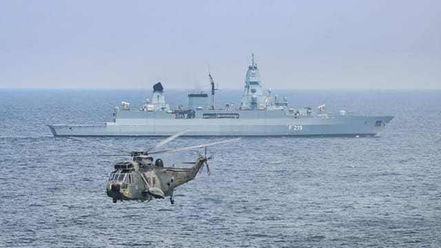 A military helicopter flies over the sea near a German warship, F219.
