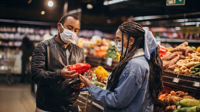 Two masked individuals selecting fresh peppers in a supermarket aisle, practicing safety during pandemic.
