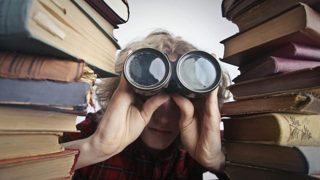 A man observing through binoculars surrounded by stacks of vintage books indoors.